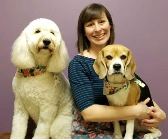 A woman smiling, seated next to a white fluffy dog and holding a beagle, both wearing patterned bandanas, against a purple background.